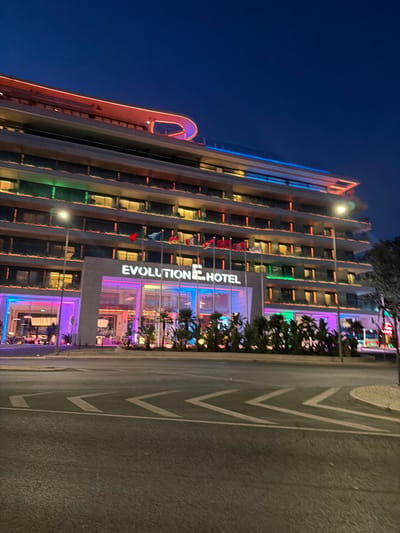 The image shows the "Evolution Hotel" in Cascais, Portugal, at dusk or night. The hotel is a multi-story building with modern architecture, illuminated by various colored lights. The main entrance is prominent, with large glass windows and colorful lighting in shades of pink, purple, and blue. Above the entrance, the hotel's name, "EVOLUTION HOTEL", is displayed in white illuminated letters.
Several flags are visible on flagpoles in front of the hotel. The building's facade features multiple floors with rows of windows, and some floors are accented with green and orange lighting. The rooftop is outlined with red and blue neon lighting.
In the foreground, a street with traffic markings is visible, suggesting the hotel is situated along a road or avenue. Palm trees and other plants are planted in front of the hotel, adding to the environment. The sky is dark blue, indicating it is nighttime or late evening. There are no discernible people in the image. The overall impression is that of a vibrant and modern hotel illuminated for the night.