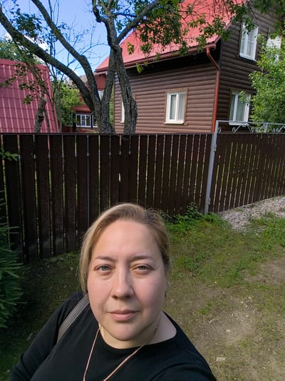 This is a selfie taken outdoors in Novonikolskoye, Russia. A Caucasian woman with blonde hair and blue eyes is in the foreground, wearing a black shirt and a gold necklace. She is looking at the camera. Behind her, a brown wooden fence separates the foreground from a residential area. Two wooden houses with red roofs are visible beyond the fence. The house on the right has a dark brown exterior with white-trimmed windows. The house on the left has a lighter brown exterior with a red metal roof. Trees with green leaves are scattered throughout the scene. The ground in the foreground is covered with grass and dirt, with some gravel on the right side. The sky is blue with some scattered clouds.