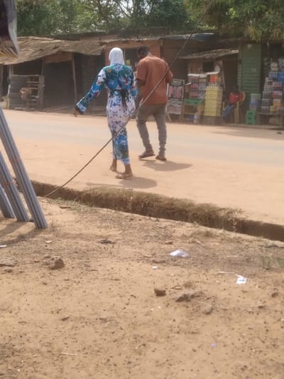 The image displays an outdoor street scene in Agege, Nigeria, under bright daylight. The foreground consists of exposed reddish-brown dirt, irregularly shaped small rocks, and scattered pieces of white debris. On the far left, a stack of grey, cylindrical pipes is positioned vertically. A thick black cable extends diagonally from the lower left quadrant towards the upper right, passing in front of two individuals.

Beyond a narrow, shallow dirt trench or culvert, two people walk away from the camera on a light-colored, unpaved road or path. The individual on the left, appearing to be female, wears a white head covering (hijab) and a full-length, long-sleeved dress with a prominent blue, white, and possibly green abstract or floral pattern. She wears light-colored flat sandals. The individual on the right, appearing male, wears an orange-brown t-shirt, grey trousers, and dark sandals or flip-flops. He holds a small, dark object in his right hand, possibly a mobile phone.

In the background, several single-story commercial structures with corrugated metal roofs line the road. The structures appear aged or in disrepair. On the left side of the background, a shop entrance shows stacked tires. On the right, another shop features a black and white "POS" (Point of Sale) sign and multiple crates containing bottled beverages (soft drinks and water) stacked outside. A person wearing a red shirt is seated on a chair further back, near the shop displaying beverages. Green foliage and trees are visible in the distant background. The overall environment appears dusty.