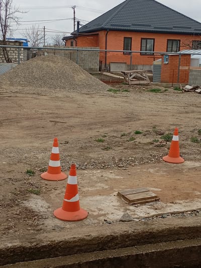 The image displays an outdoor scene in Kurganinsk, Russia, depicting an unpaved area adjacent to residential structures. The foreground features a dirt and gravel ground with a concrete trench or edge running horizontally across the bottom. Three orange and white striped traffic cones are positioned on the ground: one in the lower-left quadrant, one centrally, and one in the mid-right quadrant. A rectangular, flat wooden cover is observed on the ground to the right of the central cone. In the midground, a large, conical pile of light grey gravel dominates the left side. The background is occupied by a two-story red brick house featuring a dark grey corrugated metal roof and multiple dark-framed windows. To the immediate right of the house, a smaller, single-story brick structure with a small blue-framed window is visible. A chain-link fence, concrete block walls, and scattered construction materials are present around the property boundaries. The far background includes bare deciduous trees, utility poles with overhead lines, all under an overcast sky. No individuals are present in the frame.