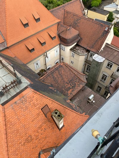 This is an aerial photograph of rooftops in the city of Bílina, Czechia. The scene is dominated by terracotta roof tiles, characteristic of European architecture. Several buildings are visible, including a prominent one with multiple dormer windows on its steeply pitched roof. Adjacent structures feature a mix of tile and darker, flat roofing materials. A modern glass-enclosed elevator shaft rises between two buildings. Details such as chimneys, gutters, and metal railings are also present. The surrounding environment shows glimpses of trees and other buildings, indicating an urban or semi-urban setting. The perspective suggests the photograph was taken from a high vantage point, possibly a taller building or tower. There are no people or animals visible in the frame.
