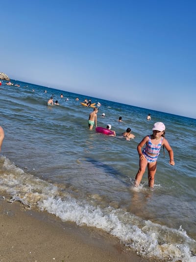 The image depicts a beach scene with numerous people enjoying the water. The primary subject in the foreground is a young girl in a pink hat and a blue and pink checkered swimsuit, kicking her legs in the shallow water, creating splashes. She appears to be having fun. Behind her, several other individuals are visible in the sea, some swimming and others standing in the water. Notable objects include a pink inflatable ring in the water.
The setting is a beach with a sandy shore and the sea. The water is a clear blue-green, with small waves breaking on the shore. The sky is a bright, cloudless blue, indicating a sunny day. The lighting suggests it is daytime, likely midday or early afternoon. The overall atmosphere seems joyful and relaxed, with people engaging in recreational activities.
The location is identified as Sveti Vlas, Bulgaria. The presence of many people enjoying the sea suggests a popular tourist destination during a warm season. No visible text is present in the image.