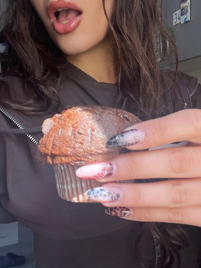 A person with long, dark hair and red lipstick is holding a chocolate cupcake. The person is wearing a black zippered jacket. The fingers holding the cupcake have long nails with various patterns and small jewels. The location is unknown, and there is no evidence to suggest it is in Groningen, Netherlands.