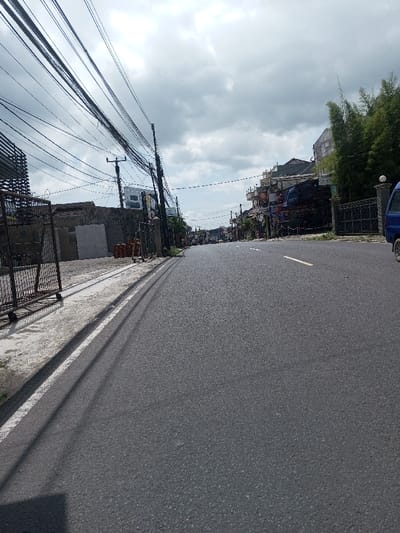 The image depicts a paved asphalt road in Gubbisamamidi, India, viewed from a low angle looking down the street. The sky above is largely overcast with scattered clouds. On the left side of the road, a concrete shoulder or sidewalk is bordered by a white line. Beyond this, a metal fence or gate stands in front of a concrete wall. Stacks of light-colored rectangular slabs, possibly construction materials, are arranged against the wall, along with smaller reddish-brown objects. Numerous utility poles line the left side, heavily laden with a dense network of black electrical and communication wires extending overhead. Further down, commercial structures are visible. On the right side of the road, a similar concrete shoulder is present. The front passenger side of a blue compact van or small truck is partially visible on the far right. Beyond this, a dark metal gate precedes a cluster of tall green bamboo plants. A row of multi-story buildings, displaying various signs and architectural details consistent with commercial or mixed-use properties, lines the street into the distance. The road itself is dark grey asphalt, appearing clear of significant debris, and a broken white lane marker is visible further down the center. No individuals are clearly identifiable as subjects within the frame.