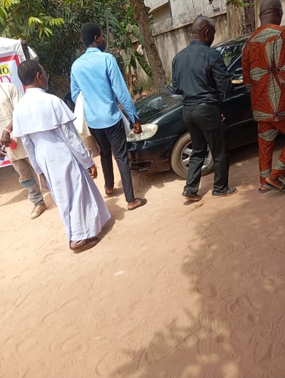 The image depicts an outdoor scene in Okpatu-Ukehe, Nigeria, under bright sunlight, with five individuals and a vehicle on an unpaved, sandy pathway.

From left to right:
1.  A partial view of an individual in a light-colored garment, holding a red object near a white banner fragment displaying "MO" and "ITY" in red text.
2.  An adult male wearing a white clerical-style robe with a distinct shoulder cape and dark brown shoes, appearing to be slightly stooped and facing towards the right.
3.  An adult male in a light blue long-sleeved button-down shirt, dark trousers, and brown sandals, facing away from the viewer, moving towards the right.
4.  An adult male in a black long-sleeved shirt, black trousers, and dark shoes, standing by the front passenger side of a dark-colored (likely black or dark green) sedan. He is also facing away from the viewer, oriented towards the right.
5.  A partial view of an adult male in an orange and brown patterned traditional Nigerian attire, with dark sandals, also facing away from the viewer towards the right.

The sedan, possibly a Toyota Corolla, is visible from its front right quarter. The ground consists of light brown, loose soil or sand, covered with multiple footprints and shoe impressions. In the background, green tropical foliage is visible alongside a light-colored building wall topped with barbed wire, and a tree trunk. The scene is brightly lit, indicating clear weather, with distinct shadows cast on the ground. The individuals appear to be part of a group, moving or standing together.