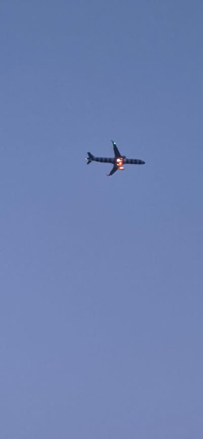 An airplane is flying in the sky. The airplane is dark with a horizontal striped pattern on its fuselage and wings. The underside of the plane's engines are glowing with orange and red light. The tail fins of the airplane are illuminated with green lights. The sky is a clear, light blue color. The image is taken from below the airplane, looking upwards. There is no visible indication of the city of Mainz, Germany within this image.