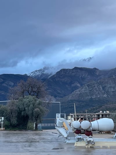 The scene depicts an outdoor industrial yard in Tomba, Montenegro, under an overcast, grey-blue sky with low-lying clouds partially obscuring distant mountain peaks. The foreground consists of a wet, dark grey paved surface. On this surface, multiple light grey industrial components are arranged. These include two white cylindrical tanks with spherical caps, and a larger white, horizontally oriented conical cement mixer drum, all mounted on light-colored frames with visible red mechanical parts beneath some components. To the left of these parts, a light grey chute-like structure is also present. In the midground, a dark grey industrial building with a wide horizontal garage-style door is visible. To its left, a cluster of green trees and shrubs grows densely. To its right, a bare deciduous tree with a complex network of branches stands. Further right and behind the machinery, a multi-story concrete skeletal structure of a building under construction is observed, alongside a wooden utility pole. The background features a range of dark, rugged mountains, with their upper slopes and peaks partially covered in white snow. No individuals, overt actions, or interactions are discernible.