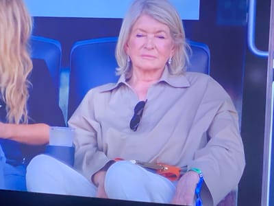 The image shows Martha Stewart sitting in a large chair, wearing a tan shirt and white pants. She is looking down and to her left, with her hands in her lap. There is a pair of sunglasses tucked into the collar of her shirt, and an orange accessory with a strap is resting on her lap. A wristband is visible on her left wrist. The background is dark and appears to be a stadium or outdoor venue. There is another person with blonde hair to her left, partially visible. The image does not provide any information about the city of Zastavna, Ukraine.
