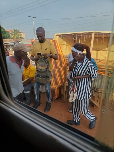 A man wearing a yellow shirt and jeans holds a dark backpack while looking at his phone. He stands to the right of an older man in a gray and red shirt and a tan cap. To the right of the man with the backpack, a woman in a black and white striped outfit, with a white headband and earbuds, walks while holding a phone and a patterned tote bag. She appears to be looking down. Behind them, a stall with a yellow tarp and red and yellow striped fabric is visible. The ground is dirt. The scene is outdoors in a city environment. The image appears to be taken from inside a vehicle looking out.