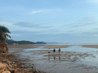 The image shows a coastal scene in Baan Sri Thanu, Thailand. A wide, shallow tidal flat dominates the foreground. The receding tide has left pools of water on the sand. Three people are walking away from the viewer on the flats. From left to right, the first person is wearing a white shirt and shorts, the second a dark top and light shorts, and the third a dark shirt and light shorts while holding a long stick. In the background, a coastline features low hills and sparse development of buildings visible along the tree line. Mountains can be seen further inland, shrouded in atmospheric haze. The sky is overcast with blue and grey clouds. Rocks and a palm tree are in the lower left corner.