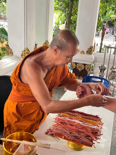 In Baan Hua Thanon, Thailand, a Buddhist monk, wearing an orange robe, is shown tying a red and white braided string bracelet around a person's wrist. The monk's head is shaved and he is looking down at his task. In front of him on a white table is a golden bowl with water and sticks, and a stand displaying a large pile of similar braided bracelets in red, yellow, and white colors. Behind the monk, pillars with ornate gold carvings and a railing are visible, with trees and other people in the background, suggesting an outdoor setting at a temple or similar religious site.