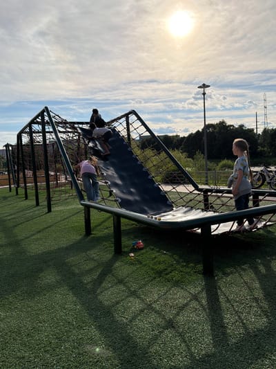 The image shows a playground with a net climbing structure. Several children are visible on and around the structure. In the foreground, a young girl in a pink shirt and blue jeans is climbing on the net. To her left, two children are crouched down on the net, one wearing a white shirt and the other a darker top. In the background, a girl with blonde hair in a ponytail, wearing a light-colored shirt with a pineapple print, stands on the net. The playground has a green, artificial turf surface. In the distance, trees and a cloudy sky with the sun are visible. A lamppost is also present. The lighting suggests it is daytime, likely late afternoon due to the angle of the sun and shadows. The overall mood appears playful and active. There is no visible text in the image.