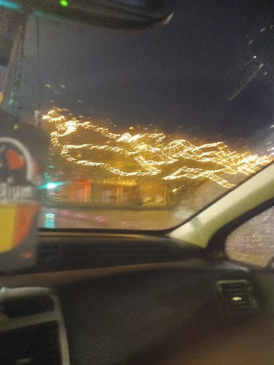 The photograph appears to have been taken from inside a car at night. A blurred, vertical display of bright, yellowish lights is visible through the car window, possibly streetlights or building lights. The window is slightly wet, causing a streaked effect. The car's interior, including part of the dashboard and a vent, is visible on the right. The overall impression is of a nighttime drive through a brightly lit area, possibly in the rain.