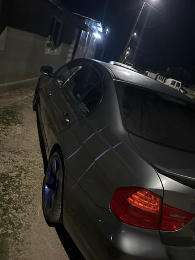The image depicts a dark grey four-door sedan, consistent with a BMW 3 Series (E90 generation), parked outdoors at night in Tsarichino, Bulgaria. The vehicle is positioned with its left side and rear quarter angled towards the viewer, occupying the central and right portions of the frame. It features dark blue multi-spoke alloy wheels, visible on both the front and rear axles on the left side. The passenger windows and rear windshield appear tinted. A small, dark-colored spoiler is affixed to the trunk lid. The vehicle's paint surface shows reflections from an artificial light source, creating distinct, angled grid-like patterns on the side body panels. The roof surface exhibits a textured or frosty appearance. The left taillight is illuminated in red.
The car is situated on an unpaved surface, likely gravel or dirt, with sparse, low-lying vegetation visible. In the background to the left, a light-colored building wall with a window is visible, along with corrugated metal siding. To the right and in the distance, a white utility van is parked. The scene is illuminated by multiple bright, artificial light sources, including a prominent streetlamp visible in the upper right corner, casting strong light and shadows. No individuals are present, and no active interactions are depicted.