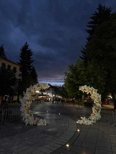 Captured at night in Razlog, Bulgaria, the image presents a street scene featuring two floral arches. The sky is a mix of dark blues and purples, suggesting twilight. Tall trees are present, some on either side of the frame. A building is visible at the bottom. The street is paved, and lighting fixtures are embedded into the pavement, illuminating the pathway. People are seen walking in the distance between the arches. Metal barriers are placed along the street. The floral arches are composed of white flowers.