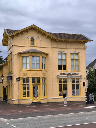 A bright yellow, two-story building with architectural detailing stands prominently on a street corner under an overcast sky. The building features a dark tiled gable roof with decorative eaves and brackets. The facade is painted a vibrant yellow, highlighting its brick texture.
On the upper floor, an arched window is centered in the gable, flanked by two decorative blind arches. A prominent bay window extends from the first floor on the corner, and multiple rectangular windows with yellow frames are visible across both floors. The ground floor consists largely of display windows and a central entrance door, all matching the building's yellow color.
Signs on the building indicate its function: "goudwisselkantoor" (gold exchange office) in white dimensional letters above the ground floor windows on the right, and a smaller blue sign with "goud wissel kantoor" on the left side. The bay window features the text "Goud Zilver Munten" (Gold Silver Coins) repeated multiple times. A small number "24" is visible on the main entrance door, and other text regarding services is displayed in one of the ground floor windows. Posters showing smiling faces are visible in two of the ground floor windows on the right.
On the sidewalk in front of the building, a young woman with blonde hair stands casually, dressed in a dark top, light pants, and sneakers, looking towards the right.
The street scene includes a brick sidewalk, a red-paved bicycle lane, and an asphalt road with white markings. A traffic light showing red is visible on the right, next to a black utility box covered in white graffiti. A black, ornate street lamp extends from the building on the left. The overall impression is a daytime scene in an urban setting, likely Alkmaar, Netherlands, as provided.