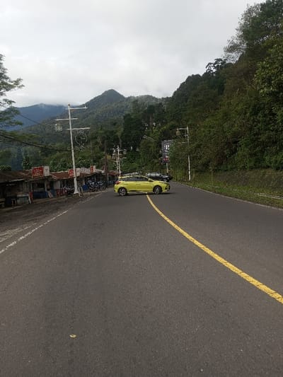 This image depicts a two-lane asphalt road, marked with a solid yellow center line curving to the right, under an overcast sky in a mountainous region, consistent with Sandamal Eliya, Sri Lanka.
On the left side of the road, a dirt shoulder borders several single-story structures with corrugated metal roofs, appearing to be roadside shops or stalls. Multiple motorcycles are parked on this shoulder in front of these structures. White utility poles with multiple cross-arms and wires are visible along this side.
On the right side of the road, a steep, densely vegetated hillside rises, covered with trees and green foliage. A dark-colored billboard is partially obscured by trees further along this side.
A light yellow/lime green hatchback vehicle is positioned on the road, angled slightly towards the left, with its front left wheel near the yellow center line, suggesting a turn or stop. A dark-colored sedan or SUV is parked further ahead on the right side of the road, partially obscured by foliage. No individuals are explicitly visible within the frame.