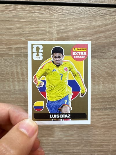 The image shows a soccer sticker featuring Luis Díaz. The sticker has a gold background and displays the FIFA logo on the left and the Panini "EXTRA STICKER" logo on the upper right. Luis Díaz is depicted in a yellow jersey with red and blue accents, and blue shorts. He is wearing the number 7. The Colombian flag is visible on the sticker. The sticker is held by a hand in the lower left corner of the image, with the person's thumb and forefinger obscuring a small portion of the sticker. The background of the image is a wooden surface. There is no information in the image that relates to the city of Prisovo, Bulgaria.