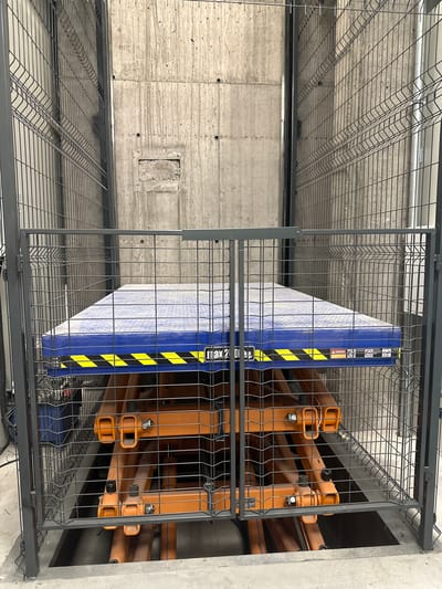 An indoor shot shows a gray metal cage surrounding a platform lift with a blue surface marked with yellow and black hazard stripes. The platform is raised by orange scissor-like supports, and a portion of the mechanism is visible below the platform. The background consists of a rough, gray concrete wall with some staining and a small rectangular opening. The cage has a dark gray frame and fine wire mesh panels. The flooring is a light gray concrete. No people are present in the image.