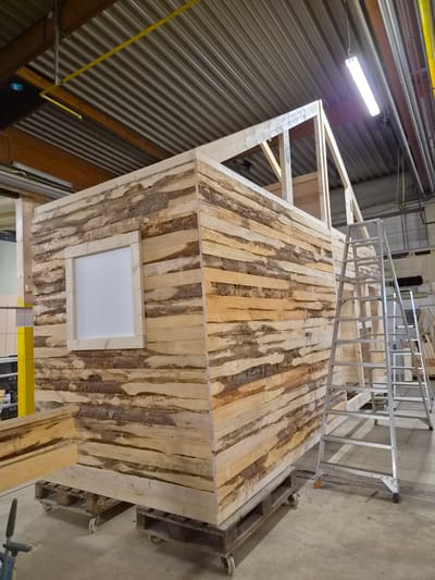 An interior shot captures a rustic wooden structure, resembling a small cabin or shed, in the process of construction within a workshop or industrial space in Leudelange, Luxembourg. The exterior walls of the structure are composed of horizontally laid, unfinished wooden planks with visible bark, giving it a rough, natural aesthetic. A small, square window with a light-colored frame is positioned on the front wall. The roof structure, made of lighter wood, is partially visible, indicating ongoing work.
To the right of the structure, a silver A-frame ladder stands upright, suggesting that work is being done at height. The floor is concrete, showing some signs of wear and tear, with a few wooden pallets scattered around, one of which supports the cabin. In the background, industrial elements are visible, including a corrugated metal ceiling with exposed beams and piping, as well as fluorescent lighting fixtures. A portion of a yellow pipe runs along the ceiling.
The scene is well-lit, primarily by the overhead lighting. There are no people visible in the image. The overall impression is one of a construction site or workshop environment where a wooden structure is being assembled.
