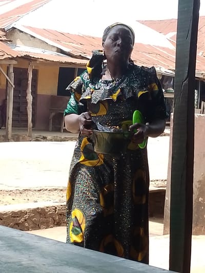 A woman stands in what appears to be a rural village setting in Iseyin, Nigeria. She is wearing a vibrantly patterned, long-sleeved dress with ruffles around the neckline and sleeves. The fabric is dark with large yellow and green circular motifs, and smaller white dots. She has a headscarf wrapped around her hair.

In her hands, she holds a shallow, dark-colored bowl filled with some items, possibly food or ingredients. To her right, she also holds a bright green, circular lid or plate. Her facial expression suggests she might be speaking or in the process of eating.

The background shows simple structures with corrugated metal roofs, suggesting a traditional or modest housing style. There are wooden posts and a stone wall, indicating an outdoor area. The lighting suggests it is daytime, with bright sunlight casting shadows. The overall scene conveys a sense of everyday life in a warm climate. No visible text is present in the image.