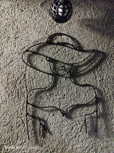 This image, taken in the city of Elin Pelin, Bulgaria, displays a highly textured, light gray stucco-like wall surface. Mounted on the wall is a black metal wire art piece depicting the stylized bust of a woman wearing a broad-brimmed hat. Above and slightly to the right of the wire art, a black spherical light fixture with a caged design encloses a white internal element. Distinct shadows are cast to the right and below both the wire art and the light fixture, indicating a strong, directional light source originating from the upper left of the frame. The bottom-left corner of the image includes a "REDMI NOTE 11 PRO 5G" watermark.