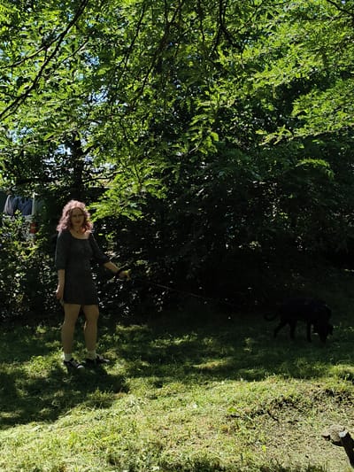 A person with reddish, curly hair, wearing glasses and a dark, short-sleeved dress with a patterned design, stands on a grassy area. The person is holding a leash, which extends to a dark-colored dog in the background. The dog appears to be sniffing the ground. The environment is outdoors, with abundant green foliage and trees casting shadows on the grass. Sunlight filters through the leaves, creating dappled patterns on the ground. In the far left background, a portion of a white vehicle is visible behind some bushes. The scene appears to be a park or garden area. The provided context indicates the location is the city of Donetsk, Ukraine.