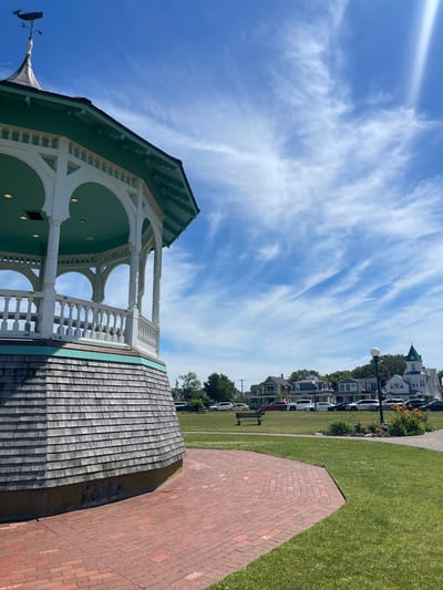 The image captures a view of a gazebo in Ocean Park, Oak Bluffs, Massachusetts, United States of America, on a bright, sunny day. The gazebo, painted in white and teal, is a prominent feature, with its wooden shingle base and ornate railings. A whale-shaped weathervane tops the structure. In the background, several Victorian-style houses line the street, typical of the Oak Bluffs architectural style. A paved brick walkway leads towards the gazebo, and a well-maintained grassy lawn surrounds it. A bench and a flower bed with blooming lilies are visible in the park. The sky is a vibrant blue with wispy white clouds, and the overall scene conveys a sense of a peaceful summer day in a charming coastal town. There are no people visible in the frame.