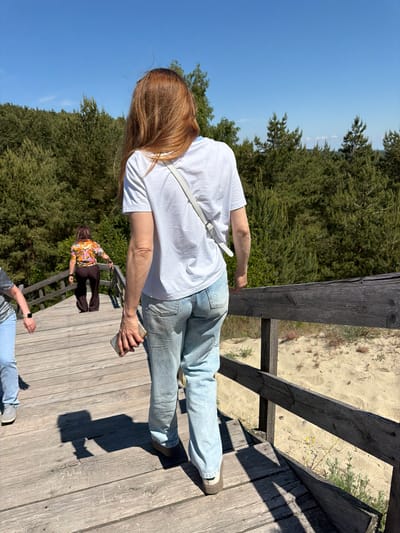 In Morskoye, Russia, a group of individuals ascend a wooden walkway situated amidst sandy dunes and dense coniferous forest. The sky is clear and blue. The foreground features a woman from the rear, with long auburn hair, wearing a white t-shirt and light blue jeans. She holds a mobile phone in her right hand. To her left, another person in light blue jeans and a grey top is partially visible. Further up the walkway, a third person, viewed from behind, is dressed in a colorful patterned shirt and dark brown flared pants. The walkway is constructed of weathered wooden planks and features a dark wooden railing. The environment suggests a natural, outdoor setting.