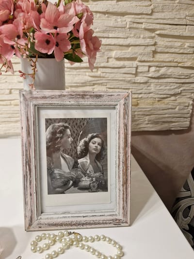 An antique framed photograph of two women is displayed on a white table. The photograph is black and white and depicts the women looking into a mirror. A vase of pink flowers sits behind the framed photograph. A pearl necklace is draped in front of the photograph on the table. The background consists of a textured, cream-colored wall. The scene is indoors, likely in a domestic setting. There is no information in the image that would suggest a specific location in Bremen, Germany.