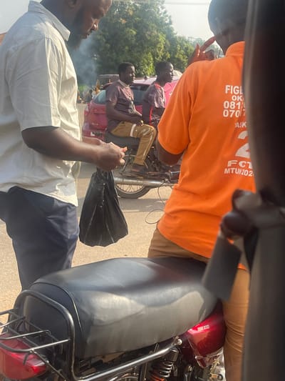 The image depicts a street scene in Galadimawa Village, Nigeria, with a focus on a group of individuals and motorcycles. In the foreground, a person wearing an orange t-shirt with white lettering, including "FIGL" and a phone number starting with "081," is partially visible from the back. They are standing next to a motorcycle with a black seat and a metallic frame. To the left, another individual in a light-colored shirt and dark pants holds a black plastic bag. In the background, two men are seated on a motorcycle. One is wearing a maroon t-shirt and brown pants, while the other is in a similar maroon t-shirt. A red vehicle is parked behind them. The environment appears to be a roadside with trees and other vehicles visible in the distance, suggesting a typical urban or semi-urban setting. Smoke is rising in the background.