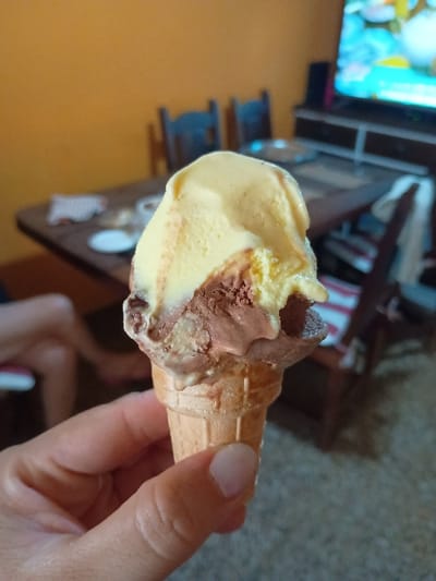 The image captures a close-up of a hand holding a melting ice cream cone indoors. The ice cream has two distinct scoops: one light yellow, likely vanilla, and another brown, resembling chocolate or mocha, both showing signs of melting and dripping down the waffle cone. A thumb and fingers with neatly trimmed, unpolished nails are visible gripping the cone.
In the blurred background, a television screen displays vibrant, colorful imagery, suggesting it's actively playing. Below the screen, there's a dark wooden or metal shelf unit. To the left, a wall painted a warm orange or peach hue is visible. Further back and out of focus, parts of what appear to be legs and feet indicate the presence of another person, possibly sitting or reclining.
The scene conveys a casual, relaxed indoor setting, possibly a living room, where someone is enjoying a sweet treat. No specific time of day or weather can be determined from the indoor shot, and there is no visible text in the image. The provided context for this media file is General Pacheco, Argentina.