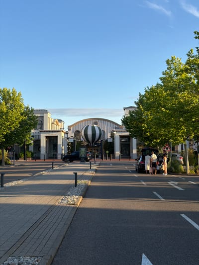 The image depicts a wide-angle outdoor view of a modern architectural complex featuring clean lines and light-colored facades, possibly a shopping outlet or a business park, situated in Ingolstadt, Germany. The sun casts long shadows across the paved pathway and asphalt road, indicating it is either early morning or late afternoon. A prominent, large hot air balloon, striped in green and white, is visible in front of one of the buildings.
To the right of the frame, a group of four individuals, two adults and two children, are gathered around the open trunk of a black SUV. One adult, a woman in a light-colored dress, appears to be loading items into the vehicle. Another adult, a man in shorts and a t-shirt, is standing nearby. The other two individuals, seemingly children, are also near the car. A second black car is parked to the right of the SUV, and a triangular road sign with a warning symbol is visible on a pole.
On the left side of the image, a tree with vibrant green foliage shades a portion of the pathway. The pathway itself is paved with interlocking bricks and bordered by a strip of small white pebbles and dark grey curbs. Several black bollards are spaced along the edge of the pathway.
The sky is clear and blue, with a few wispy clouds visible in the upper portion of the image. The overall scene suggests a pleasant day in a well-maintained urban or suburban environment. The specific location is identified as the Ingolstadt Village, a luxury outlet shopping center.