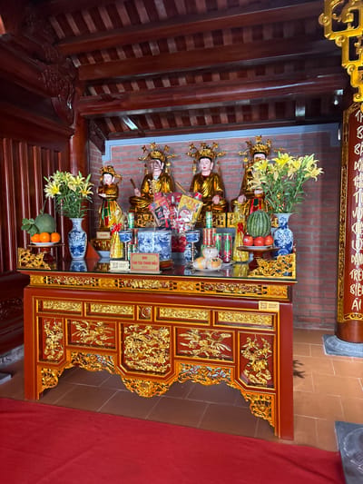 The image depicts an altar inside a building, likely a temple or shrine, located in Diên Khánh, Vietnam. The altar is elaborately decorated in red and gold, with intricate carvings. On the altar, there are offerings, including fruit, flowers, and small statues of deities. These statues are adorned with golden crowns and robes. Ceramic vases with blue and white patterns are also present. The background reveals a brick wall and wooden beams of the building's structure. A red cloth hangs near the altar.