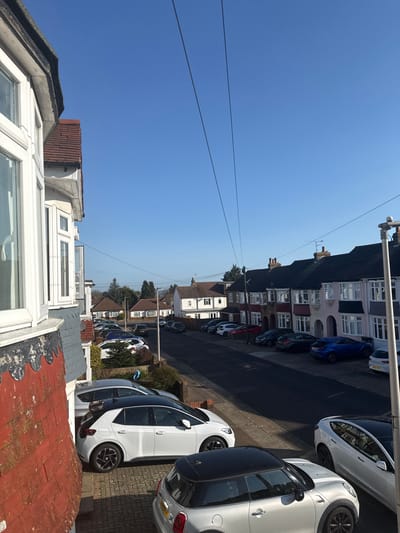 The image displays a sunlit residential street in Rochester, United Kingdom, viewed from an elevated vantage point, likely an upper-floor window. The sky is clear and bright blue, intersected by two prominent dark utility wires stretching diagonally across the upper portion of the frame.
In the immediate foreground on the left, a section of a building facade is visible, characterized by red brickwork, decorative grey scalloped slate tiles along the lower edge, and white framed bay windows. Below, on a paved driveway, a white Volkswagen ID.3 electric vehicle is parked, partially obscured by a silver Mini Cooper with a black roof, its UK license plate visible at the bottom right.
The street extends into the midground, appearing to gently curve to the left, and is densely lined with numerous parked cars on both sides. The residences along the street are predominantly semi-detached or terraced, featuring varied facades, bay windows, pitched tiled roofs, and chimneys. A grey street light pole is present on the far right edge of the frame. The scene is illuminated by strong direct sunlight, creating distinct shadows. No individuals are visible.
