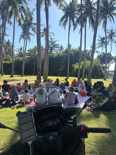 The image depicts a group of people picnicking on a grassy area under a grove of tall palm trees, likely in Karangasem, Indonesia, given the tropical vegetation and the presence of a Yamaha scooter in the foreground, which may be a mode of transport for locals. The people are mostly seated on blankets, facing each other, suggesting a social gathering. They are dressed in casual attire, with some individuals wearing headscarves. The atmosphere appears relaxed and communal. The scooter's handlebars and part of its front are visible, with a smartphone mounted and displaying a map application. Sunlight filters through the palm fronds, casting dappled shadows on the ground. In the background, more trees and some indistinct structures can be seen. The overall scene is one of outdoor leisure and community interaction in a tropical setting.