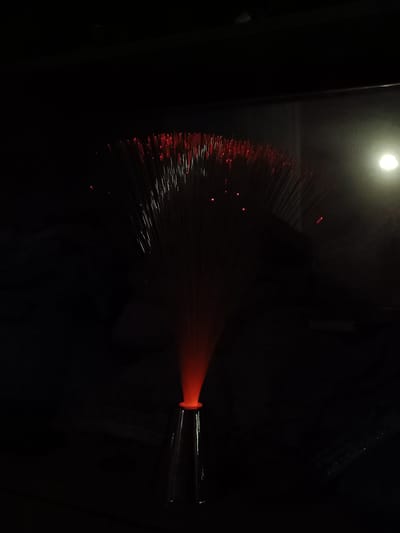 A dimly lit photograph shows a fiber optic lamp. The lamp has a base that flares out at the top. The fibers extend upwards and outward, with small points of red light visible at the tips of many fibers. The background is dark and indistinct, but a faint light source is visible in the upper right. The location in Cherkovitsa, Bulgaria is not discernible from the image.