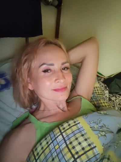 A woman with blonde hair is lying in bed, looking at the camera. She is wearing a green tank top and has makeup on, including eyeshadow and lipstick. Her left arm is behind her head, and she is partially covered by a blanket with a plaid and floral pattern. The room appears to be dimly lit, and there is a black cloth hanging on the wall behind her. The image is a selfie taken indoors, presumably in a bedroom. There is no visible information in the image to indicate a specific location within Shalsky, Russia.
