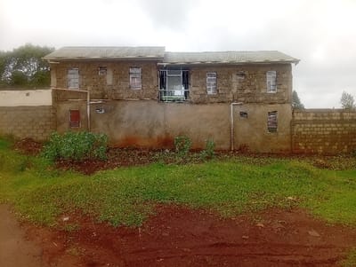 A two-story, grey building with a corrugated metal roof sits behind a low brick wall. The lower section is plastered, while the upper floor features exposed blocks with windows. A small red door is visible on the left. Vegetation fills the space between the building and the foreground. The ground is a mix of red soil and green grass. The sky is overcast.