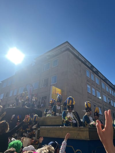 The image shows a procession of people in traditional costumes, likely participating in a parade or carnival in Cologne, Germany. The event is taking place in the city center, close to the intersection of Schildergasse and Hohe Straße. The people are standing on a large, ornate float adorned with gold decorations and a blue canopy. Many of them are wearing blue and gold uniforms, reminiscent of historical military attire. Some are holding flowers or other decorations, while others are waving to the crowds. The scene is captured from a low angle, looking up at the float and the people on it. The bright sun is visible in the clear blue sky, illuminating the facade of a large building with multiple windows, likely a residential or commercial building on Hohe Straße.  The overall atmosphere is festive and celebratory, with the bright colors and cheerful faces reflecting the joy of the occasion.  A hand, likely belonging to a spectator, is reaching up towards the float.