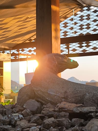 The image captures a statue of a Komodo dragon in Labuan Bajo, Indonesia, set against a backdrop of a sunset. The statue is positioned in the foreground, partially obscured by a pile of rough, grey rocks. It is made of a textured, dark material, likely concrete or a similar composite, mimicking the scales of a real Komodo dragon. The statue is large and appears to be a permanent fixture.
Behind the statue, a wooden beam and a section of a building's roof with a patterned ceiling are visible. The roof has a distinct slatted design that casts striped shadows across its surface, illuminated by the warm orange light of the setting sun. Beyond this structure, a diamond-patterned screen or fence is visible, with the sky beyond.
The lighting suggests late afternoon or early evening. The sun, low on the horizon, casts a bright glare to the right of the statue's head, creating lens flare effects and illuminating the scene with a golden hue. In the distance, hills or mountains are silhouetted against the pale sky, indicating a coastal or island environment characteristic of Labuan Bajo.
The overall impression is one of an outdoor setting, possibly a resort, a public park, or a viewpoint, where the Komodo dragon statue serves as a prominent decorative or cultural element. The presence of lush green foliage and a glimpse of what appears to be a seating area or terrace in the background further suggests a developed area designed for visitors. No people are visible in the image.