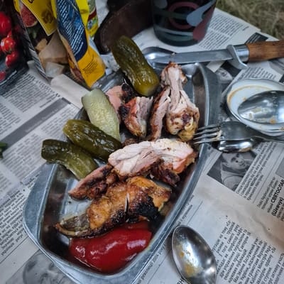 The image depicts a picnic scene laid out on newspapers. In the foreground, a metal plate holds a meal consisting of grilled meat, several pickled cucumbers, and a dollop of red sauce, likely ketchup. A fork rests on the plate, partially embedded in the meat. To the right of the plate, a metal spoon with some food residue is visible. In the background, a knife with a wooden handle is placed on the newspapers, and a cup with a patterned design is also present. The newspapers themselves appear to be in Ukrainian. The overall impression is a casual outdoor meal, possibly a picnic. The mention of Chmyrivka, Ukraine, is not directly inferable from the visual information in the image itself, as there are no identifying landmarks or text within the image that specify this location.