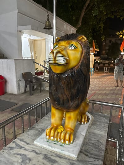 The image depicts a statue of a lion, positioned prominently near the entrance of a temple in Vadodara, India. The lion statue is painted yellow and brown. A small arrangement of yellow flowers rests on one of its paws. The statue sits atop a white, rectangular base, placed on a light gray, marble-patterned surface. A metal railing is visible in the background, suggesting the statue is located at the top of a staircase. In the background, people can be seen walking in the distance. A bell hangs from the temple structure.