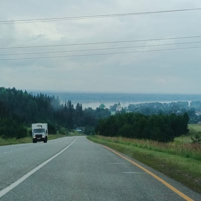 The image depicts a scenic view of a road leading towards a distant landscape, likely in the vicinity of Нечкино, Russia. The foreground is dominated by a grey asphalt road with white and yellow lane markings. A white box truck is visible driving away from the viewer on the left side of the road. The middle ground features a dense, dark green forest that lines the right side of the road and extends into the distance. Beyond the forest, a body of water is visible, likely a river or lake, with a hazy horizon. On the far side of the water, there are more trees and what appear to be buildings, including a prominent structure with green domes, possibly a church or monastery. The sky is overcast and pale blue, with several thin, dark horizontal lines across it, suggesting power lines. The overall atmosphere is calm and somewhat subdued due to the cloudy weather.