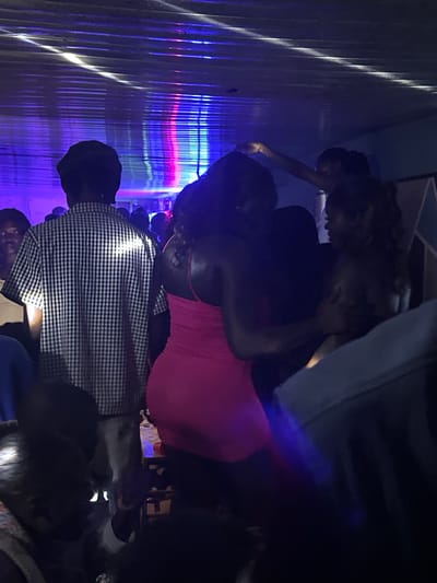 In Wumat, Nigeria, a dimly lit indoor space with a corrugated metal ceiling is illuminated by colored stage lights, casting blue and pink hues. Several individuals are visible, mostly obscured by darkness or the angles of the shot. In the foreground left, a person's head is partially visible, facing down. To their right, a person wearing a black and white checkered short-sleeved shirt is seen from the back, with a dark cap on their head. In the center, a woman in a bright pink, form-fitting dress is embraced by another individual whose face is not clearly visible. To the right of them, another person's face is partially visible in profile, looking away. The scene appears to be a gathering or event, possibly a party or nightclub, characterized by low lighting and dancing or socializing.