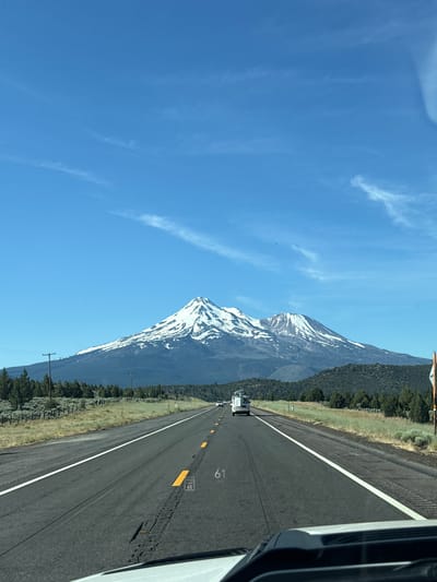 The image depicts a view from a vehicle traveling on a highway. The highway runs parallel to a large, snow-capped mountain. The mountain is covered in patches of snow, contrasting with the dark rock and green vegetation on its lower slopes. A dense forest of trees flanks the mountain and extends towards the roadside. The sky is a clear, bright blue with a few wispy white clouds.

The road appears to be a multi-lane highway with a white dividing line. Several yellow markers are visible along the side of the road, some with numbers. One marker clearly shows the number "65", and another further down the road has "61" visible. A white truck is seen in the distance, traveling in the same direction as the viewer. The right side of the image shows the interior of a vehicle, suggesting the photo was taken from the passenger seat or driver's side.

The scene is set during the daytime, likely late morning or early afternoon, given the bright sunlight and clear sky. The weather appears to be pleasant, with sunshine and a lack of precipitation. The presence of snow on the mountain suggests a location at a higher altitude or a cooler climate. The highway and the surrounding landscape indicate a rural or semi-rural setting. The overall impression is one of a scenic drive through a mountainous region. There is no indication of people in the image, apart from the implied presence of the photographer and any occupants of the vehicle.