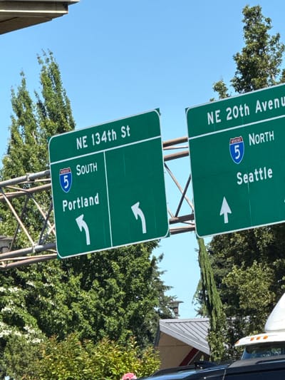 The image shows a road sign for Interstate 5 in Vancouver, Washington, United States of America. The sign indicates directions for NE 134th St South to Portland and NE 20th Avenue North to Seattle. The sky is clear blue, and lush green trees are visible in the background. A portion of a vehicle is also visible in the foreground.