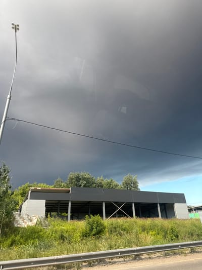 The image depicts a construction site for a building with a dark grey metal facade and exposed steel beams. The structure appears to be partially covered with white and camouflage-patterned tarps. It is surrounded by tall green grass and trees. In the background, there is a dark, stormy sky, contrasted with a patch of blue sky on the right. A tall lamppost with multiple lights is visible on the left side of the image, with a black wire extending horizontally across the frame. A metal guardrail runs along the bottom of the image, separating the construction site from what appears to be a road or pathway. The scene is located in Yam, Russia. There are no people visible in the image.