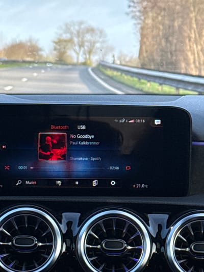 The scene depicts the interior of a vehicle, specifically the dashboard and infotainment system, viewed from the driver's perspective, with the road ahead visible through the windshield. The vehicle is located in Puurs, Belgium.

The upper portion of the image shows a paved, two-lane road curving slightly to the right, under a hazy or overcast sky. On the right side of the road, a metal guardrail runs alongside a vegetated embankment with bare trees. Similar trees are visible in the distance on the left side of the road. White dashed line markings are visible on the road surface.

The central part of the image features a black digital infotainment screen displaying music playback details. The screen indicates "Bluetooth" and "USB" connectivity. The current time is 08:16, with a strong cellular signal and a message icon present. The song playing is "No Goodbye" by "Paul Kalkbrenner," originating from "Shamakova - Spotify." The track is 00:01 seconds into playback, with 02:46 remaining. Below the track information, control icons for "Muziek" (Music), playback controls (play/pause), and settings are visible. The internal temperature displayed is 21.0°C.

The lower portion of the image shows three round, chrome-accented air conditioning vents, typical of a modern Mercedes-Benz vehicle, below the infotainment screen. No individuals are visible in the frame, and no explicit interactions are occurring beyond the automated music playback. The environment suggests an open road or rural area on a clear day with some atmospheric haze.