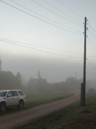 The image depicts a foggy morning scene in the village of Bolshaya Usa, Russia. A white SUV is parked on a dirt road in the foreground, to the left of the frame. The road curves into the distance, disappearing into the thick fog. On the right side of the road, a wooden utility pole with wires stretches across the frame. In the background, silhouetted against the fog, are a few houses and trees. The overall atmosphere is serene and slightly melancholic, characteristic of a misty rural morning.